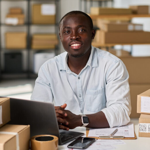 Happy young owner of small warehouse looking at camera by his workplace with gadgets, documents and stacks of packed boxes
