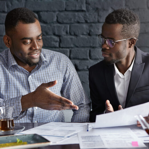 Attractive African businessman in glasses and suit holding papers, showing presentation of his project to cheerful business partner, who smiling happily, supporting his idea with positive gesture